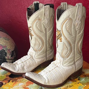 Könnte beinhalten: Ein Paar cremeweiße Cowboystiefel mit einer strukturierten, schuppenartigen Oberfläche am Fußteil und einem glatten Schaft mit goldfarbenen Stickereien, darunter ein kleines Alligator-Motiv an der Seite und braune Blockabsätze.