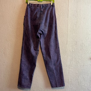 May include: Purple acid wash jeans hanging on a green hanger. The jeans have a high waist and a straight leg. The denim is a faded purple color with a textured appearance. The back pockets and belt loops are visible.