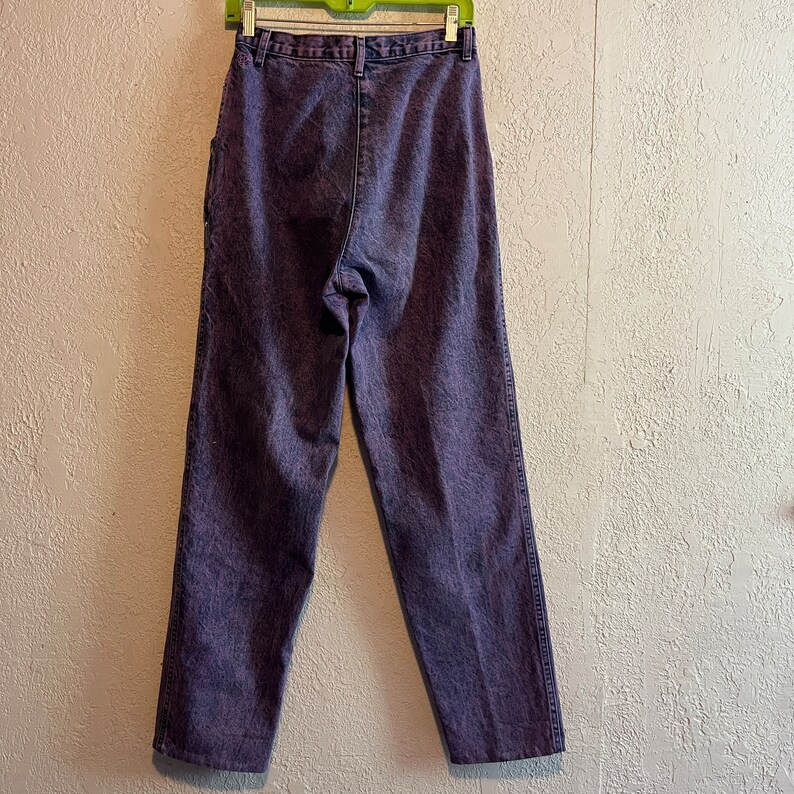 May include: Purple acid wash jeans hanging on a green hanger. The jeans have a high waist and a straight leg. The denim is a faded purple color with a textured appearance. The back pockets and belt loops are visible.