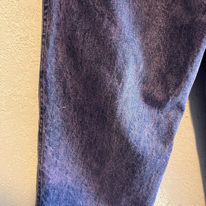 May include: Close-up of a pair of denim jeans with a purple and blue acid wash effect. The jeans have a classic five-pocket design and a straight leg cut. The fabric has a textured appearance.