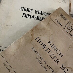 Puede incluir: Dos manuales de papel antiguos con cubiertas de color blanco roto y manchas de agua de color marrón oscuro. El manual superior dice 8-INCH HOWITZER M2 (MOUNTED IN COMBAT VEHICLES) RESTRICTED DEPARTMENT OF THE ARMY. El inferior dice ATOMIC WEAPONS EMPLOYMENT.