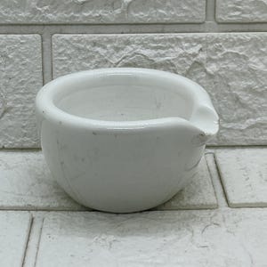 May include: A white ceramic mortar with a smooth, rounded bowl shape and a small integrated pouring spout on the right side. The interior and exterior surfaces are finished in a glossy white glaze, showing a simple, functional design.