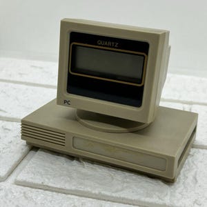 May include: A beige and brown desktop clock shaped like a vintage computer. The clock face is digital and displays "QUARTZ" on the top and "PC" on the bottom.