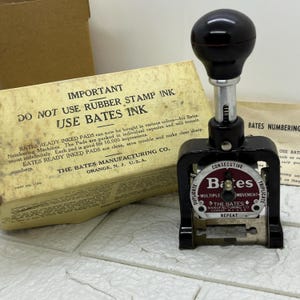 May include: A black Bates numbering machine with a round black handle and silver stem. The machine has a circular dial with the brand name "Bates" and text that reads "Duplicate, Consecutive, Multiple Movement, Triplicate, Repeat". A box with text is in the background.