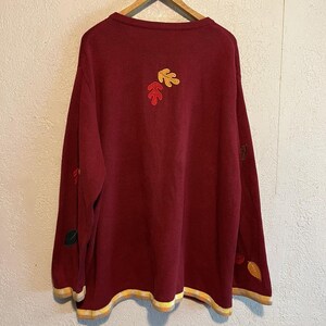 May include: A burgundy sweater with embroidered autumn leaves in shades of red, yellow, and green. The sweater has a ribbed knit texture, long sleeves, and a round neckline. The hem and cuffs are trimmed with a yellow and cream striped pattern.