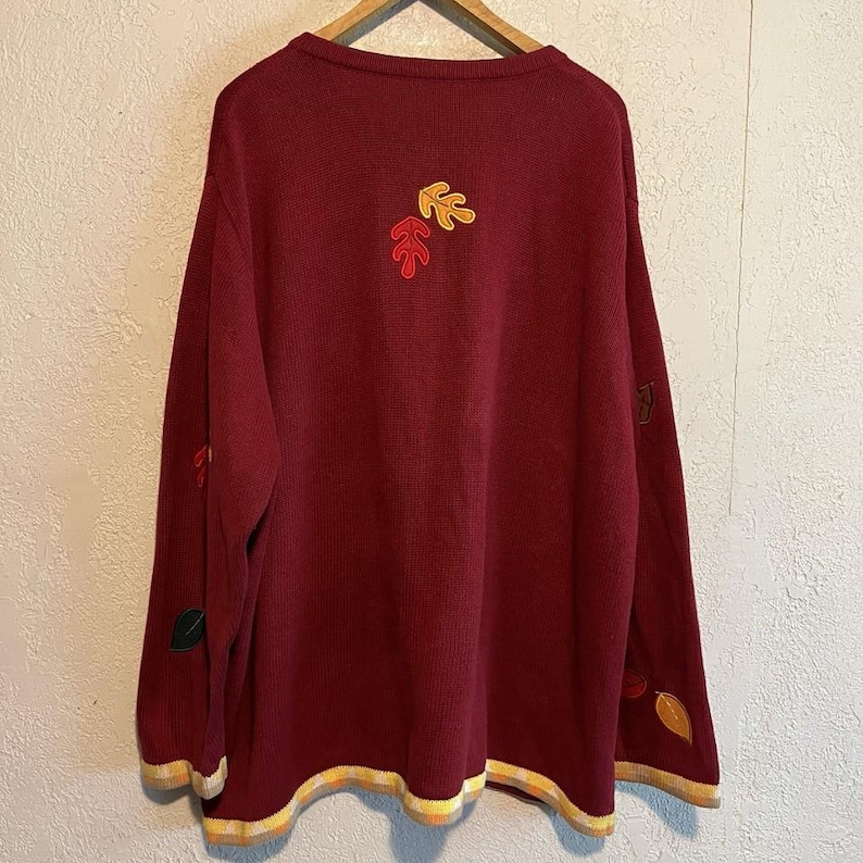 May include: A burgundy sweater with embroidered autumn leaves in shades of red, yellow, and green. The sweater has a ribbed knit texture, long sleeves, and a round neckline. The hem and cuffs are trimmed with a yellow and cream striped pattern.