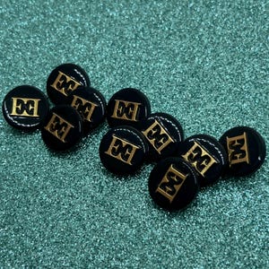 May include: A collection of ten round black buttons, each featuring a raised gold-colored emblem in the center. The emblem consists of the letters E and G intertwined in a stylized font. The buttons have a smooth, glossy finish and a circular shape.