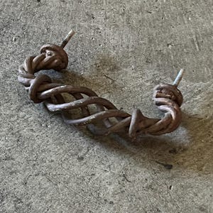 May include: A rustic, brown metal handle with a twisted rope design. The handle has two threaded posts for mounting. The hardware is likely for a cabinet, drawer, or other furniture piece.