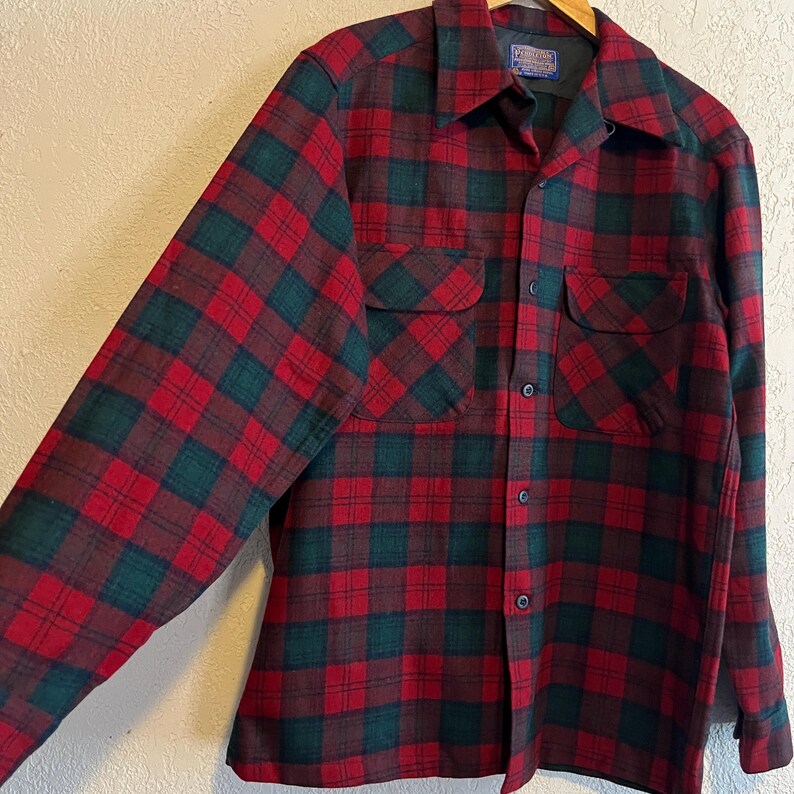 May include: A long-sleeve button-up shirt featuring a red and dark green plaid pattern. The shirt includes a pointed collar, two chest pockets with buttoned flaps, and a front button closure. A Pendleton brand label is visible at the inner neck.