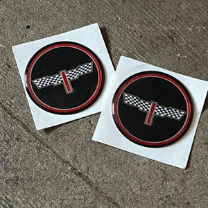 May include: Two round black and red stickers with a checkered flag design. The stickers have a red border and a central emblem featuring a checkered pattern and a vertical red stripe.