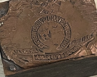 Puede incluir: Un bloque de impresión de cobre rectangular vintage con texto e imágenes en relieve. El bloque presenta un diseño circular central con texto, rodeado por una pancarta y elementos decorativos. El bloque está montado sobre una base de madera oscura y asegurado con pasadores metálicos.