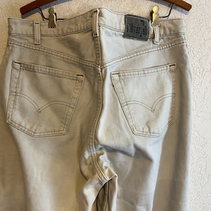 May include: Light beige denim jeans featuring a high-rise waist, two back pockets with arched stitching, and a dark grey leather-like patch on the waistband with the text silverTab printed in black. The garment includes belt loops and seams.