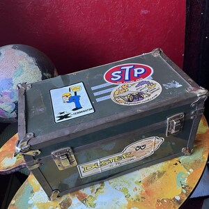May include: A rectangular olive green metal storage box with reinforced metal corners and two front latches. The lid features an STP sticker, a Road Runner sticker with the text Beep Beep, and a Terminator sticker. The front displays a Duster by Plymouth sticker.