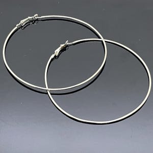 May include: A pair of large, thin, circular silver-toned hoop earrings. Each earring features a smooth, continuous wire design with a hinged clasp mechanism at the top for secure closure. The earrings have a polished, reflective metallic finish.