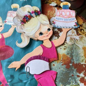 Könnte beinhalten: Eine Vintage-Geburtstagskarte mit einer Papierpuppe Ballerina, die einen rosa-weißen Kuchen mit den Worten "Happy Birthday" hält. Die Ballerina hat blonde Haare, ein rosa Kleid und eine Blumenkrone.