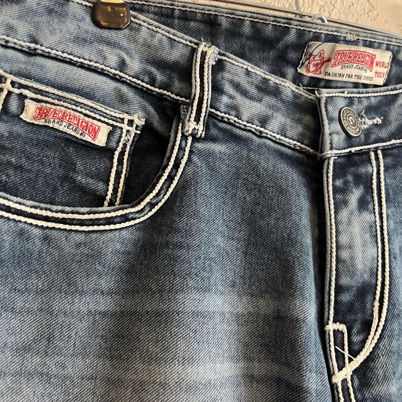 Pu&ograve; includere: Primo piano di jeans blu con cuciture bianche. I jeans presentano un classico design a cinque tasche, una chiusura con bottone e un'etichetta del marchio. I dettagli delle cuciture evidenziano le tasche e le cuciture. I jeans hanno un lavaggio sbiadito.