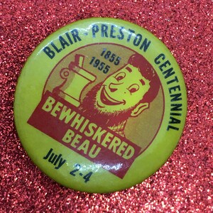 Op de afbeelding: Gele knop met een cartoonman met een rode baard en snor. De knop zegt "Blair Preston Centennial 1855 1955 Bewhiskered Beau July 2-4".