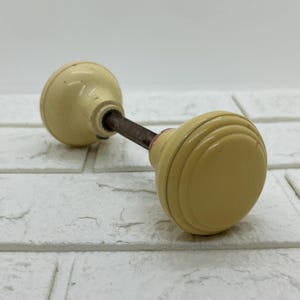 May include: A pair of cream-colored round door knobs connected by a central metal spindle. Each knob features a smooth, circular face with concentric ring detailing around the edges, showing a light, uniform finish and a sturdy, functional design.