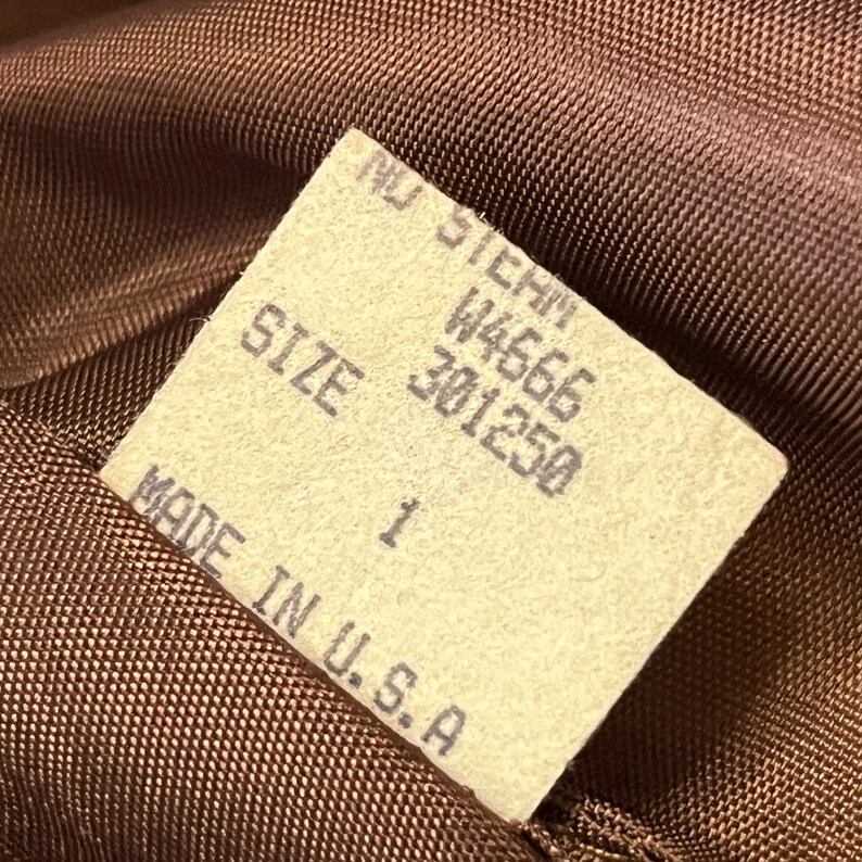 Puede incluir: Una etiqueta de prenda de tela marr&oacute;n con texto impreso en negro. La etiqueta muestra el texto NU STEHM, W4666, 301250, SIZE 1 y MADE IN U.S.A. La etiqueta est&aacute; unida a un tejido marr&oacute;n, texturizado y tejido con un patr&oacute;n de rejilla visible.