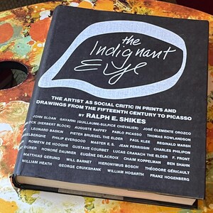May include: A hardcover book titled "The Indignant Eye" with a black cover and white text. The book's title is in a stylized font, and the cover also lists the author and a list of artists.