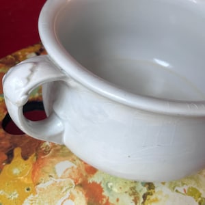 May include: Close-up of a white ceramic bowl with a handle. The bowl has a rounded shape and a glossy finish. The handle is intricately designed. The bowl is placed on a colorful surface with red, orange, and yellow hues.
