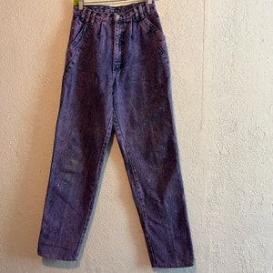 May include: Purple acid wash jeans with a high waist and classic five-pocket design. The denim pants are hanging on a hanger against a neutral-colored wall. The jeans have a button and zipper closure.