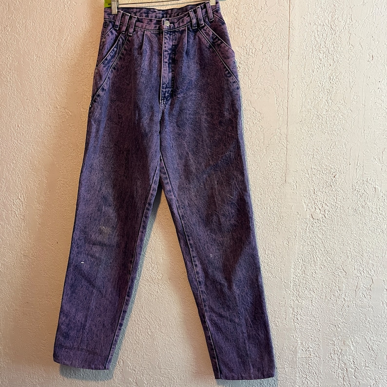 May include: Purple acid wash jeans with a high waist and classic five-pocket design. The denim pants are hanging on a hanger against a neutral-colored wall. The jeans have a button and zipper closure.