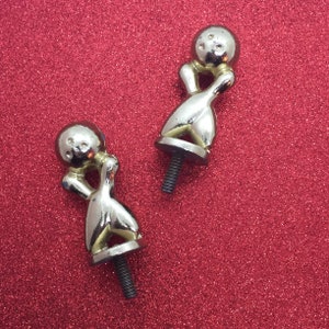 Może przedstawiać: Dwa srebrne trofea w kształcie kręgli z piłką do kręgli na górze. Trofea znajdują się na czerwonym tle z brokatem.