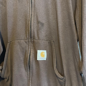 May include: A brown zip-up hoodie with a kangaroo pocket and the Carhartt logo patch. The hoodie has a full zipper and a hood. The fabric appears to be a medium-weight material, suitable for cooler weather.