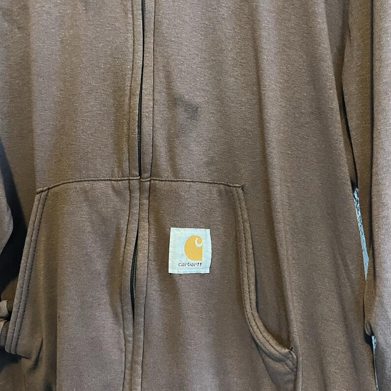 May include: A brown zip-up hoodie with a kangaroo pocket and the Carhartt logo patch. The hoodie has a full zipper and a hood. The fabric appears to be a medium-weight material, suitable for cooler weather.