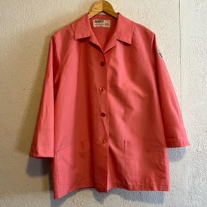 May include: A coral-colored button-down jacket with a collared neckline, three-quarter length sleeves, and two large front patch pockets. The garment features a central row of matching buttons and a small white brand label inside the collar.