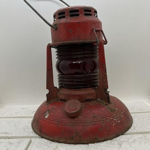 May include: Vintage red lantern with a handle and a red glass lens. The lantern has a round base and a cylindrical body with ventilation slots at the top. The metal shows signs of wear and rust, giving it a rustic appearance.