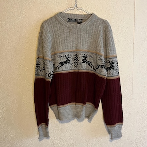 May include: A gray, burgundy, and black sweater with a reindeer and tree pattern. The sweater has a ribbed knit design on the upper and lower sections, with a cable knit pattern in the middle. The sleeves and cuffs are gray.
