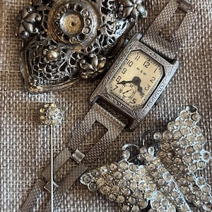 Puede incluir: Una variedad de joyas vintage, que incluyen un reloj de pulsera plateado con una esfera rectangular y una correa estampada. También, un broche plateado, un alfiler y un broche de mariposa con pedrería.
