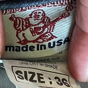 Pu&ograve; includere: Primo piano di un'etichetta di abbigliamento con la scritta "made in USA" e la taglia "36". L'etichetta presenta una grafica rossa di una persona. &Egrave; visibile anche la scritta "UNB DRY IN SHADE".