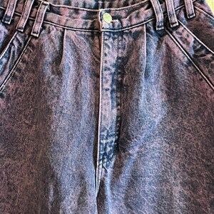 May include: Close-up of acid wash denim jeans in shades of blue and purple. The jeans have a button closure, belt loops, and pleats at the waist. The fabric has a textured appearance.