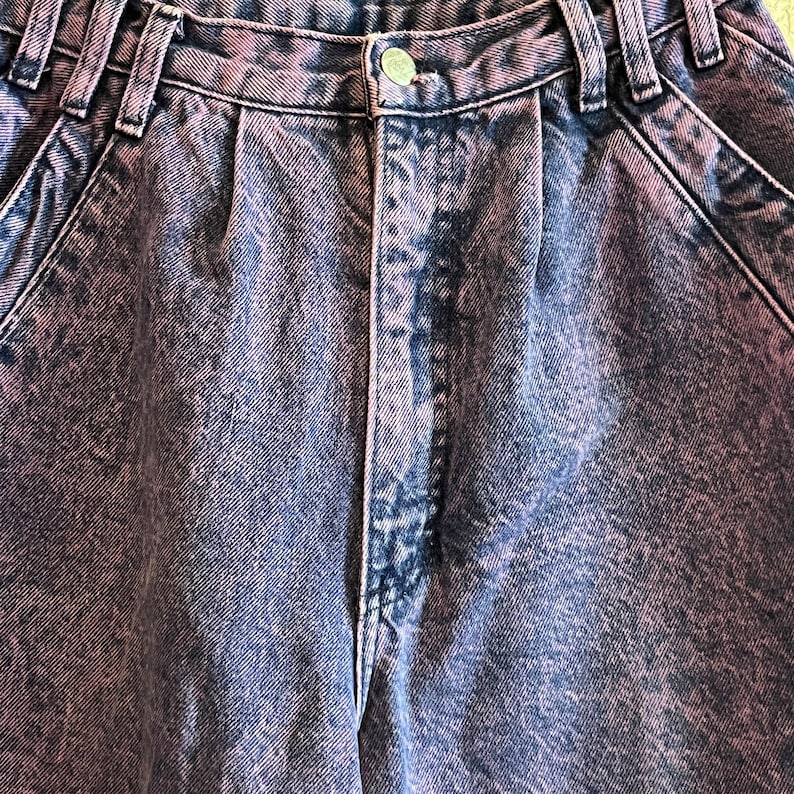 May include: Close-up of acid wash denim jeans in shades of blue and purple. The jeans have a button closure, belt loops, and pleats at the waist. The fabric has a textured appearance.