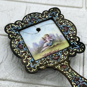 May include: A hand mirror with an ornate, dark metal frame featuring intricate blue, gold, and purple enamel filigree. The central square panel is made of white ceramic and depicts a painted cherub in a landscape, showing a visible crack.