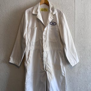 May include: Off-white work jumpsuit with long sleeves, a collared neckline, and multiple pockets. A blue oval patch with the name "Louie" is on the chest. The jumpsuit is hanging on a wooden hanger.