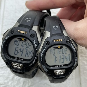 Puede incluir: Dos relojes digitales Timex Ironman Triathlon negros. Los relojes muestran la hora y la fecha en una pantalla digital. El reloj tiene una correa negra y un bisel plateado. El texto "TIMEX" y "IRONMAN TRIATHLON" son visibles. Resistente al agua hasta 100 metros.