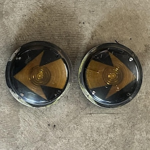 May include: Two round, vintage amber-colored vehicle lights with a black and gold arrow design. The lights have a clear, circular lens and a black metal rim. The lights are likely for a car or truck.