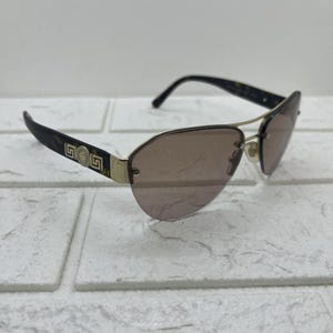 May include: Aviator-style sunglasses with brown tinted lenses and gold-tone metal frames. The temples are dark brown with a tortoiseshell pattern and gold accents. The sunglasses are set against a white brick background.