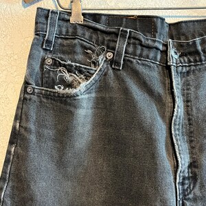 K&ouml;nnte beinhalten: Nahaufnahme einer schwarzen Jeans. Die Jeans hat eine abgenutzte Tasche mit ausgefransten Kanten und Metallnieten. Der Stoff weist Gebrauchsspuren und Ausbleichungen auf, was auf eine Vintage- oder Used-&Auml;sthetik hindeutet.