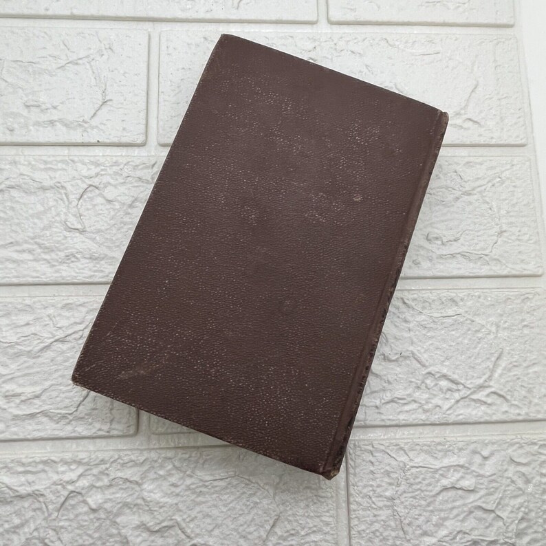 May include: A rectangular book with a solid dark brown cover featuring a textured, pebbled surface. The book is positioned at an angle, displaying its front cover and a portion of the spine, with a simple, unadorned design and no visible text.