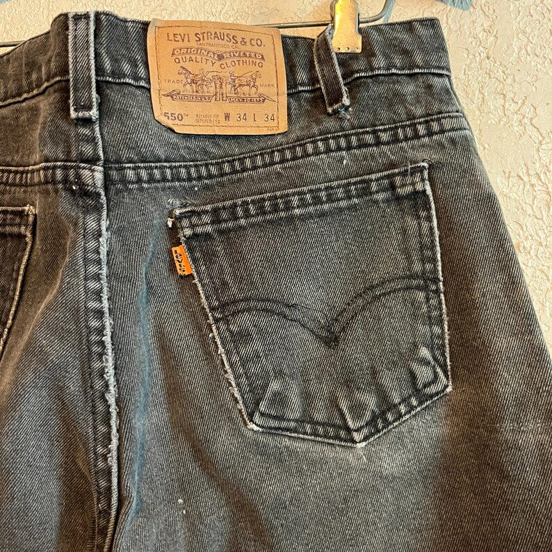 K&ouml;nnte beinhalten: Nahaufnahme einer verblichenen schwarzen Levi Strauss 550 Jeans. Die Jeans hat ein klassisches F&uuml;nf-Taschen-Design mit einem abgenutzten Look. Die Ges&auml;&szlig;tasche zeigt das Levi's-Logo und ein orangefarbenes Etikett. Taillengr&ouml;&szlig;e W34 L34.