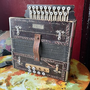 May include: Vintage accordion with a dark brown wooden body, metal details, and a leather strap. The accordion has a keyboard with white keys and the text "HEINRICH SUHR BEVERAGE, WASH." on a small label. The instrument is placed on a colourful table.