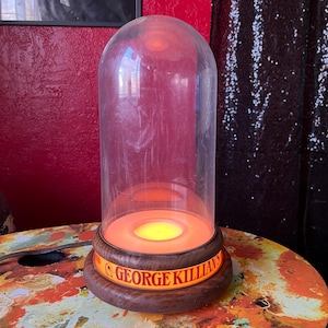 May include: A vintage cloche lamp with a clear, dome-shaped glass cover. The lamp has a wooden base with the text "GEORGE KILLIAN'S" in orange lettering. The interior glows with an orange and yellow light, creating a warm ambiance. The lamp is on a colorful, patterned surface.