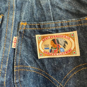 May include: Close-up of blue denim jeans with a Levi Strauss & Co. patch on the back pocket. The patch features an illustration of a Native American chief and the text "QUALITY CLOTHING."