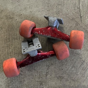 May include: Two skateboard trucks with red metal components and orange wheels. The trucks have a silver-colored base and show signs of wear. The wheels are a faded orange color.