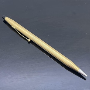 Könnte beinhalten: Ein goldfarbener Stift mit einem strukturierten, geriffelten Körper und einer silbernen Spitze. Der Stift hat einen goldenen Clip und eine schwarze Spitze. Der Stift ist auf einer reflektierenden schwarzen Oberfläche abgewinkelt.